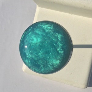 May include: A round, teal-colored knob with a shimmering, marbled effect. The knob is set against a white background, suggesting a furniture or cabinet hardware component. The knob's diameter appears to be approximately 1.5 inches.