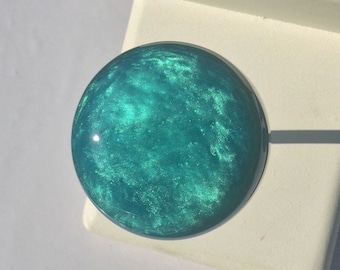 May include: A round, teal-colored knob with a shimmering, marbled effect. The knob is set against a white background, suggesting a furniture or cabinet hardware component. The knob's diameter appears to be approximately 1.5 inches.