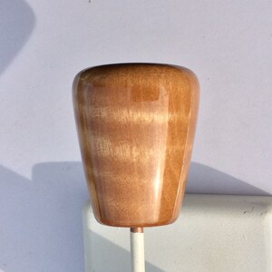 May include: A wooden knob with a glossy finish, featuring a light brown color with visible wood grain. The knob is mounted on a white metal rod. The knob's shape is a rounded, inverted cone.
