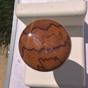 May include: A circular knob featuring a warm brown wood grain pattern with dark, irregular, meandering lines across the surface. The knob is mounted on a smooth, off-white, rectangular base with a curved top edge and a flat, solid front face.