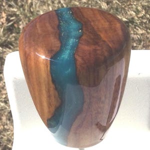 May include: A handcrafted wooden shift knob with a glossy finish. The knob features a river of teal-colored resin flowing through the center, creating a unique design. The wood has a rich, brown color.