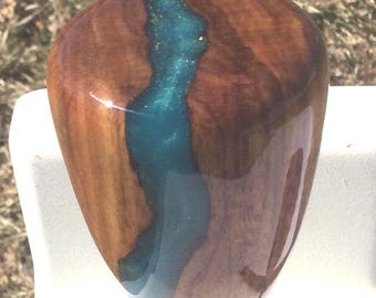May include: A handcrafted wooden shift knob with a glossy finish. The knob features a river of teal-colored resin flowing through the center, creating a unique design. The wood has a rich, brown color.