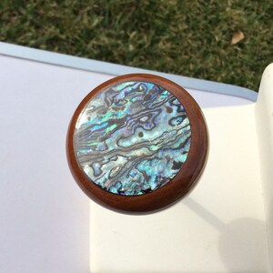 May include: Round wooden bottle stopper with a shimmering abalone shell inlay. The shell features iridescent blues, greens, and purples, set within a dark brown wooden frame. A decorative item for wine bottles.