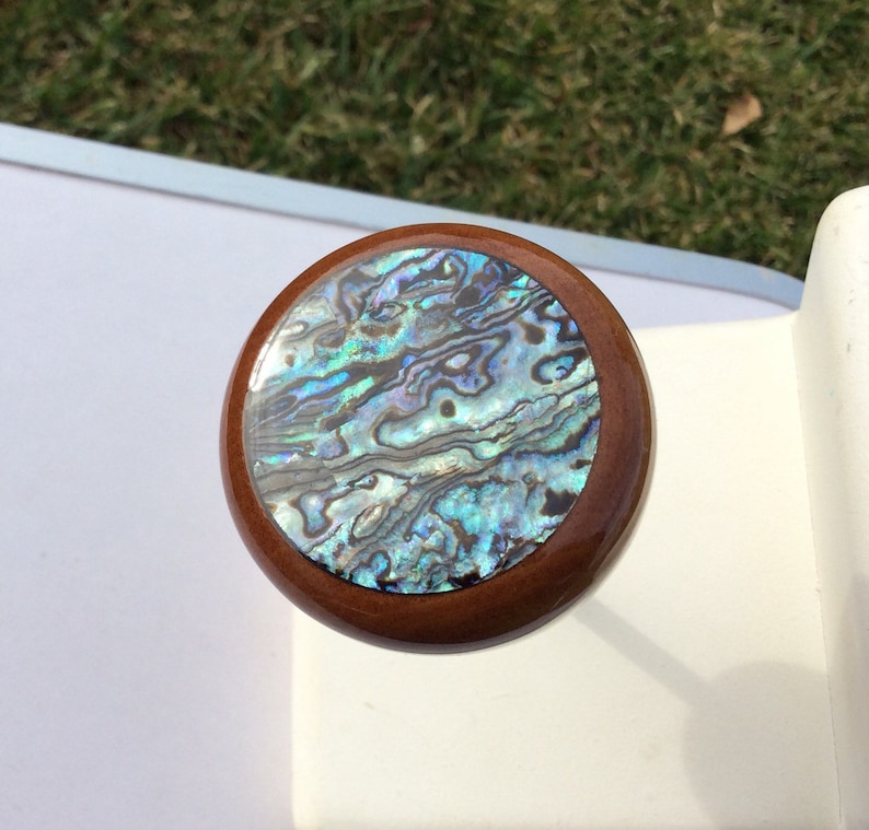 May include: Round wooden bottle stopper with a shimmering abalone shell inlay. The shell features iridescent blues, greens, and purples, set within a dark brown wooden frame. A decorative item for wine bottles.