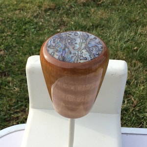 May include: A wooden bottle stopper with a polished finish. The top features an abalone shell inlay with iridescent blues, greens, and purples. The stopper is mounted on a white metal stem.