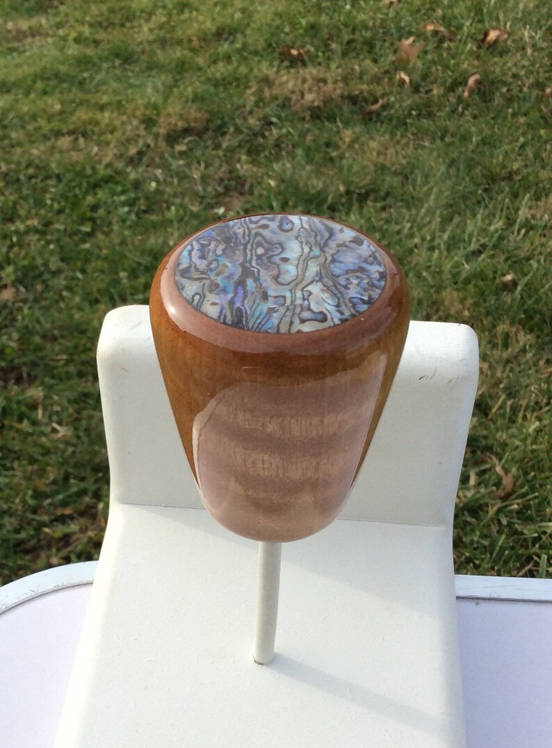 May include: A wooden bottle stopper with a polished finish. The top features an abalone shell inlay with iridescent blues, greens, and purples. The stopper is mounted on a white metal stem.