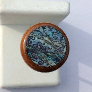 May include: A round wooden knob with a polished, dark brown finish. The center features a swirling pattern of iridescent blues, greens, and purples, resembling abalone shell. The knob is likely for a cabinet or drawer.