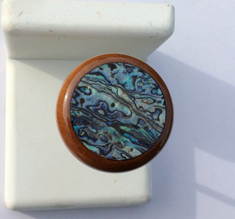 May include: A round wooden knob with a polished, dark brown finish. The center features a swirling pattern of iridescent blues, greens, and purples, resembling abalone shell. The knob is likely for a cabinet or drawer.