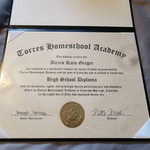 May include: A high school diploma from Torres Homeschool Academy issued to Alexis Rain Geiger in Colorado Springs, Colorado, on July 8, 2026. The document features black text on cream paper, a gold embossed seal, and two signatures in a black folder.