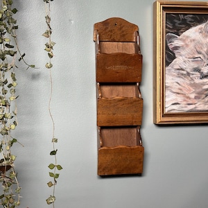 May include: A vertical wall-mounted wooden letter organiser featuring three tiered storage pockets. The top pocket is embossed with the word LETTERS in a decorative script font. The unit is constructed from medium-toned brown wood with scalloped edges.