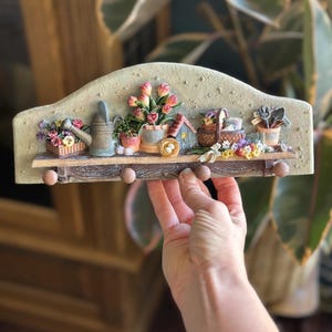 Peut inclure: Une étagère murale décorative avec un panneau arrière beige texturé en forme d'arche et une base en bois brun à quatre crochets arrondis. L'étagère présente des objets de jardin miniatures dont un arrosoir, des tulipes, un nichoir et des outils.