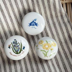 Peut inclure: Trois boutons de placard en céramique blanche avec des motifs floraux. L'un présente des fleurs bleues, un autre des fleurs jaunes et le troisième une illustration d'Hyacinthus Orientalis avec des papillons.