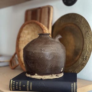 May include: A brown ceramic pitcher with a spout sits on a navy blue book with gold lettering that reads "Bread Into Roses" by Kathleen Norris and "Collier".