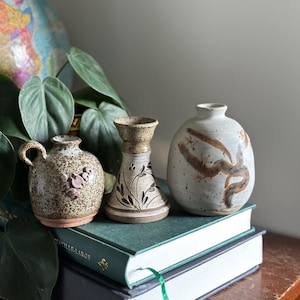 Peut inclure: Trois vases en céramique faits à la main de formes et tailles variées. Les vases sont dans des tons de marron et de blanc, avec des surfaces texturées et des motifs décoratifs. Ils sont posés sur une pile de livres verts, avec une plante feuillue et un globe en arrière-plan.