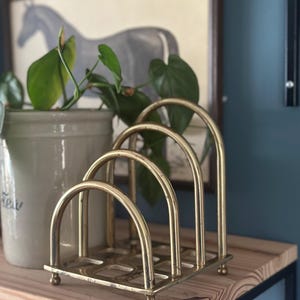 May include: A brass letter holder featuring four graduated arched dividers attached to a square base with small ball feet. The metal has a polished finish with visible signs of wear and patina, creating a structured organiser for mail or documents.