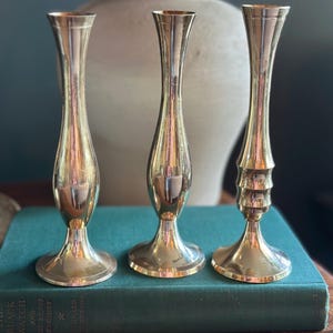 Peut inclure: Trois bougeoirs dorés de forme élancée et allongée. Chaque bougeoir a une base évasée et un sommet étroit, avec de légères variations dans la forme du corps. Ils sont disposés sur un livre de couleur sarcelle, avec un vase de ton neutre en arrière-plan.