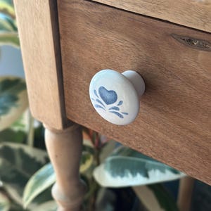 May include: A close-up of a wooden drawer with a white ceramic knob. The knob features a blue heart design. The drawer is part of a light brown wooden table with a turned leg. Green plants are visible in the background.