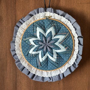 Peut inclure: Une décoration murale circulaire en tissu avec un motif d'étoile pliée dans des tons bleu sarcelle, gris et blanc. Le centre est encadré par un cercle en bois, entouré d'une bordure en dentelle blanche et d'un volant en tissu gris plissé.