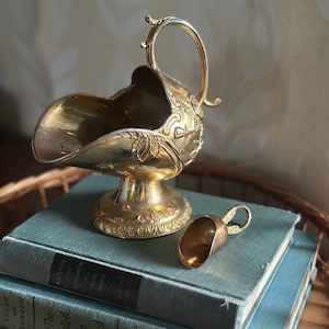 May include: A gold-coloured sugar bowl with a matching scoop, resting on two teal-coloured books. The sugar bowl has a handle and ornate floral detailing. The scoop is small and has a loop handle. The items are on a woven tray.