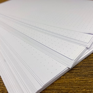 Könnte beinhalten: Ein Stapel weißer Dot-Grid-Papierbögen, aufgefächert, um die Kanten zu zeigen. Das Papier hat ein subtiles Dot-Grid-Muster und ist ideal für Bullet-Journaling, Notizen oder Skizzen. Das Papier liegt auf einer Holzoberfläche.