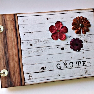 Könnte beinhalten: Ein weißes Gästebuch aus Holz mit braunem Holzrand. Das Buch ist mit drei Stoffblumen in Rot-, Orange- und Lilatönen verziert. Das Wort "GÄSTE" ist auf der Vorderseite des Buches gedruckt.
