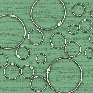 Könnte beinhalten: Eine Sammlung silberner Metallringe in verschiedenen Größen, angeordnet auf einer grün strukturierten Oberfläche. Die Ringe sind alle kreisförmig und haben eine glatte, polierte Oberfläche.
