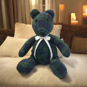 May include: A plush teddy bear with a green and blue tartan pattern and a white satin ribbon tied around its neck. The bear is sitting on a light brown surface.