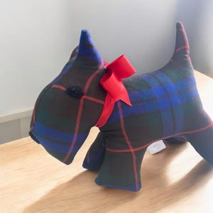 Peut inclure: Un chien en peluche en forme de terrier écossais. Le chien est fait de tissu écossais noir, bleu et rouge et a un nœud rouge autour du cou.