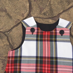 Puede incluir: Un vestido sin mangas de cuadros blancos y rojos con botones negros. El vestido tiene un cuello negro y un ribete blanco alrededor del escote.