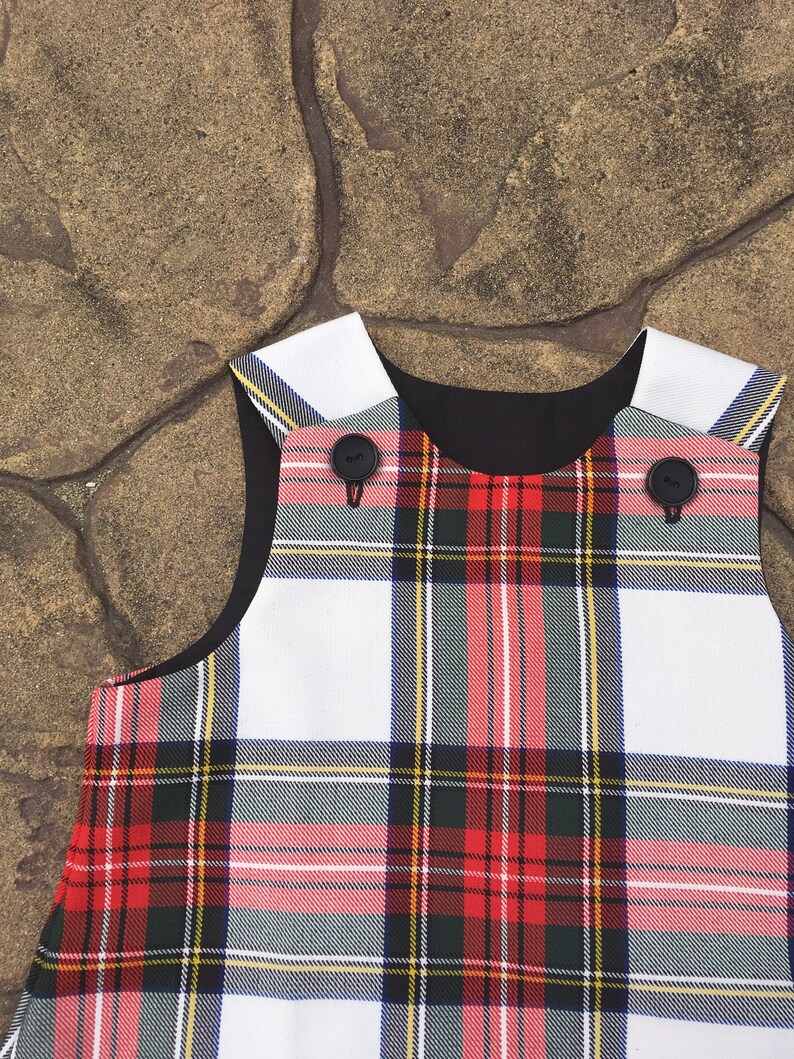 Puede incluir: Un vestido sin mangas de cuadros blancos y rojos con botones negros. El vestido tiene un cuello negro y un ribete blanco alrededor del escote.