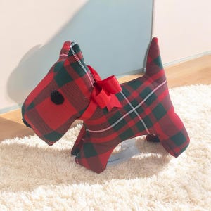 Può includere: Giocattolo di peluche a forma di cane in tessuto scozzese rosso e verde con un fiocco rosso. Il giocattolo è seduto su una superficie bianca e soffice.