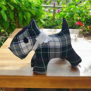 May include: A plush toy dog in the shape of a Scottish Terrier. The dog is made of green, white, and blue plaid fabric and has a white bow tie. The dog is sitting on a wooden table.