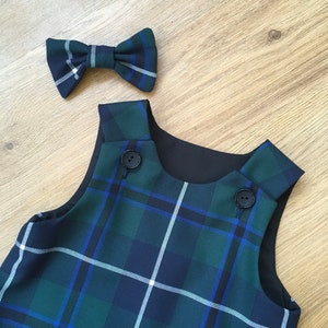 Peut inclure: Une robe chasuble à carreaux verts, bleus et blancs avec un nœud papillon assorti. La robe est sans manches, avec des boutons noirs et une doublure noire. Le nœud papillon reprend le motif à carreaux. Présenté sur une surface en bois brun clair.