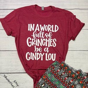 May include: A burgundy t-shirt with the text "IN A WORLD full of GRINCHES be a CINDY LOU" in white. Paired with gray pajama pants featuring a festive pattern of penguins, Christmas trees, and snowflakes. The Etsy shop name "WhatinTheCraftMan" is visible.