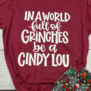 May include: Burgundy t-shirt with the text "IN A WORLD full of GRINCHES be a CINDY LOU" in white. The shirt is paired with gray pajama pants patterned with Christmas trees, gifts, and Santa figures.