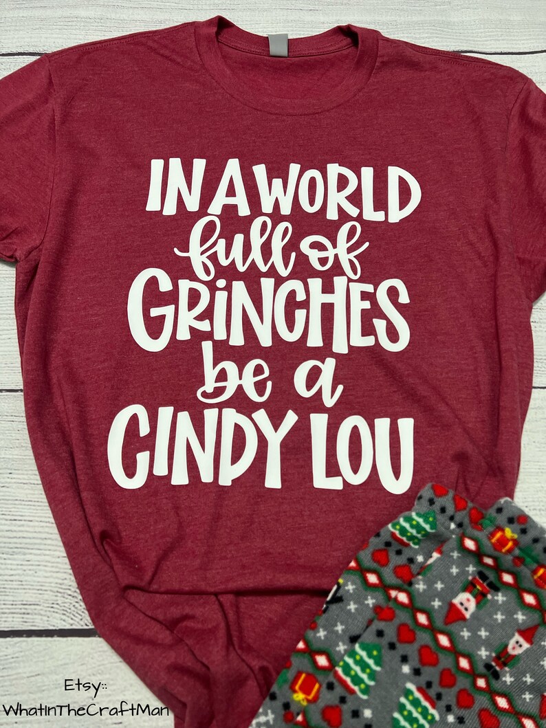 May include: Burgundy t-shirt with the text "IN A WORLD full of GRINCHES be a CINDY LOU" in white. The shirt is paired with gray pajama pants patterned with Christmas trees, gifts, and Santa figures.