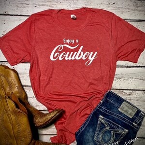 May include: Red t-shirt with the text "Enjoy a Cowboy" in white script. The shirt is paired with denim shorts and brown cowboy boots. The boots have intricate stitching and a textured pattern.