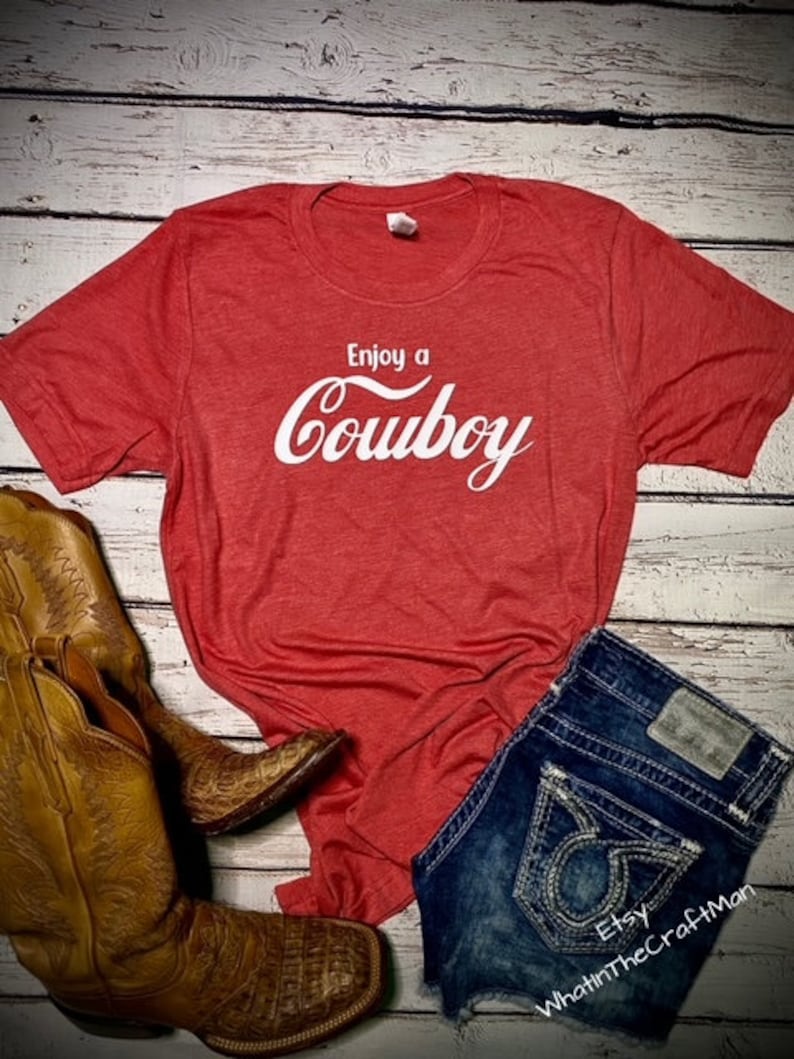May include: Red t-shirt with the text "Enjoy a Cowboy" in white script. The shirt is paired with denim shorts and brown cowboy boots. The boots have intricate stitching and a textured pattern.