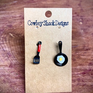 May include: A pair of earrings featuring a black frying pan with a yellow egg and a black spatula with a red handle. The earrings are displayed on a brown card with the text "Cowboy Shack Designs".
