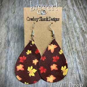 May include: A pair of brown leather earrings with a fall leaf pattern. The earrings are teardrop shaped and have silver hooks. The earrings are 2 1/4 inches long and 1 1/2 inches wide. "These are a tad bigger!" and "Cowboy Shack Designs" are written on the card behind the earrings.
