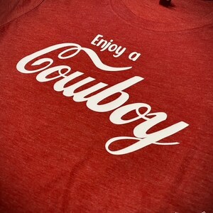 May include: A red t-shirt with the words "Enjoy a Cowboy" in white script. The shirt has a crew neck and a tag on the collar. The Etsy shop name "WhatintheCraftMan" is visible in the bottom right corner.