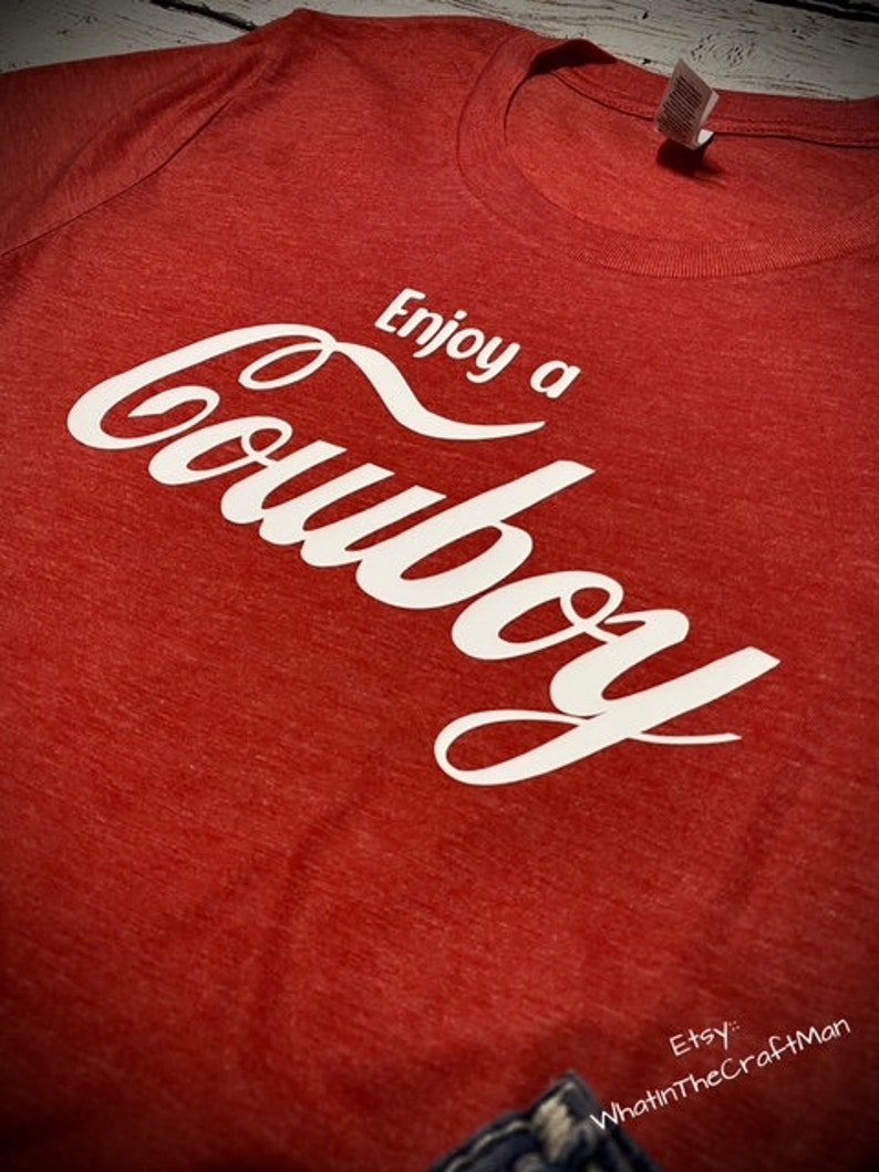 May include: A red t-shirt with the words "Enjoy a Cowboy" in white script. The shirt has a crew neck and a tag on the collar. The Etsy shop name "WhatintheCraftMan" is visible in the bottom right corner.