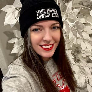 May include: A woman wearing a black beanie with the text "Make America Cowboy Again" in white. She is wearing a grey sweatshirt with a red candy cane design and the text "Old Fashioned Candy Cane Hand Rolled Since 1947".