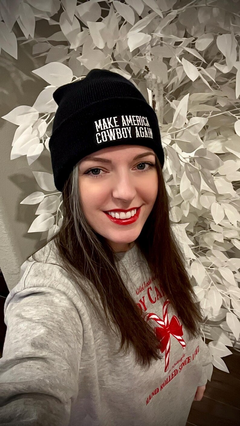May include: A woman wearing a black beanie with the text "Make America Cowboy Again" in white. She is wearing a grey sweatshirt with a red candy cane design and the text "Old Fashioned Candy Cane Hand Rolled Since 1947".