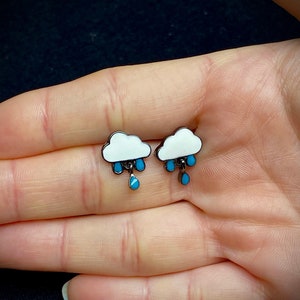 May include: A pair of silver earrings with a white cloud design and blue raindrops hanging from the bottom.
