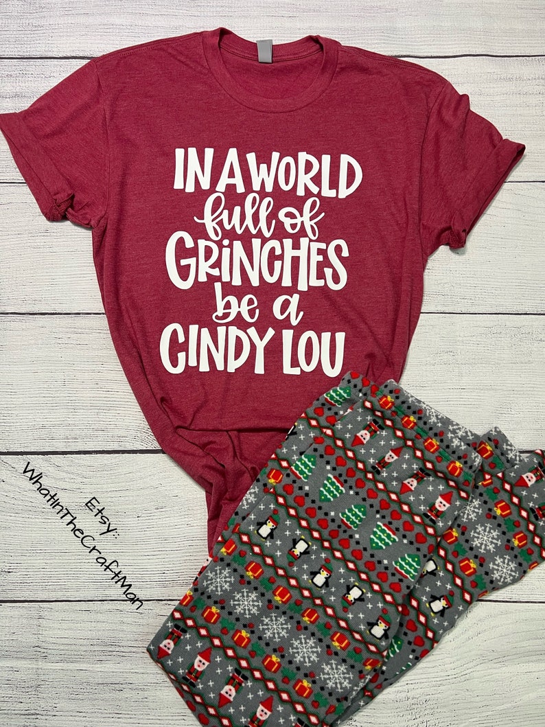 May include: A burgundy t-shirt with the text "IN A WORLD full of GRINCHES be a CINDY LOU" in white. Paired with gray pajama pants featuring a festive pattern of penguins, Christmas trees, and snowflakes. The Etsy shop name "WhatinTheCraftMan" is visible.