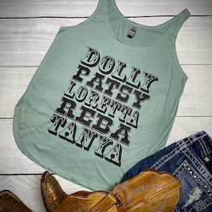 May include: Sage green tank top with black text that reads "DOLLY PATSY LORETTA REBA TANYA". The tank top is laying on a light wood surface with a pair of brown cowboy boots and blue denim shorts.