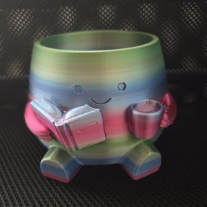 Puede incluir: Una maceta multicolor impresa en 3D con forma de personaje de dibujos animados, sosteniendo un libro y una taza. La maceta es una mezcla de azul, verde, rosa y amarillo. El personaje tiene una cara sonriente y está sentado.