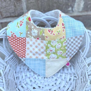 Pode incluir: Babete de bebé em patchwork em forma de coração, com tecidos estampados florais e geométricos em tons pastel. O babete tem uma borda branca e está colocado numa cesta de vime branca.