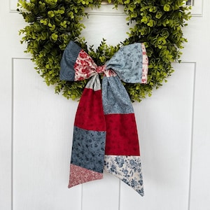 May include: A decorative fabric bow featuring a patchwork design of red, blue, and cream floral and paisley patterns. The bow has two long, hanging tails and is centered on a circular wreath made of small, vibrant green leaves.